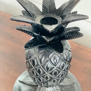 May include: A dark brown, carved wooden pineapple candle holder. The pineapple has a textured surface with a detailed leaf crown and a round base. The candle holder is designed to hold a single candle.