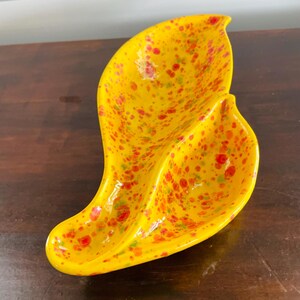 May include: A yellow, leaf-shaped ceramic dish with red and green speckled accents. The dish has a glossy finish and a unique, organic design. This decorative piece is ideal for serving snacks or as a decorative accent.