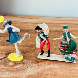 Pode incluir: Três bonecas de lã feitas à mão vestidas com roupas coloridas. As bonecas estão em pé sobre uma superfície de madeira. Uma boneca está segurando uma pá amarela, outra está usando um traje verde e vermelho, e a terceira está usando um vestido verde com uma borda vermelha e branca.