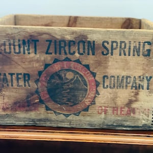 Pode incluir: Uma caixa de madeira vintage com o texto "Mount Zircon Spring Water Company" impresso na frente. A caixa tem um logotipo circular vermelho e preto com uma montanha e um sol.