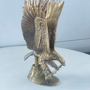 May include: A detailed brass eagle sculpture with outstretched wings, perched on a small base. The eagle holds a branch in its beak, showcasing intricate feather detailing and a golden hue. This decorative piece is ideal for home decor.