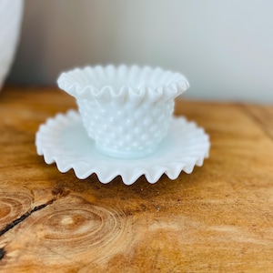 May include: A white hobnail glass candle holder with a ruffled rim, sitting on a matching saucer. The candle holder has a textured, dotted surface. The set is displayed on a wooden surface.