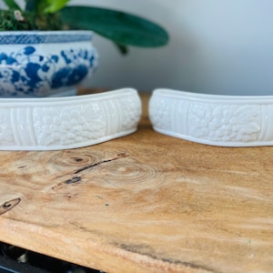 May include: Two white ceramic decorative bowls with a raised floral design. The bowls are on a wooden surface.