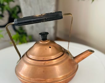 May include: A copper teapot with a black wooden handle and a brass spout. The teapot has a round body and a domed lid.