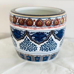 Puede incluir: Una maceta de cerámica con base blanca y patrones intrincados en azul y dorado. El borde superior muestra una banda de motivos de pétalos rojos y dorados, mientras que el cuerpo presenta formas de nubes azules y volutas ornamentales repetidas.