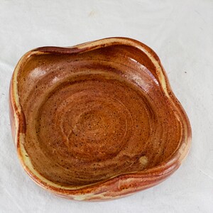Puede incluir: Un pequeño plato de cerámica de forma cuadrada con un borde ondulado. El plato está esmaltado en color marrón y amarillo con un acabado moteado.