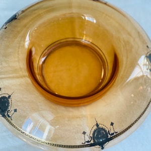 Puede incluir: Cuenco de vidrio color ámbar con un borde ancho y una zona central ligeramente hundida. El borde presenta un patrón decorativo negro y dorado con elementos geométricos y florales estilizados. El cuenco es probablemente vintage.