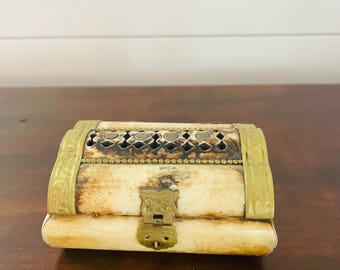 May include: A small, rectangular box with a cream-colored body and brass accents. The top features a decorative, openwork design. The box has a brass clasp and hinges. The box is likely used for storing small items.
