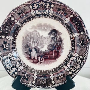 May include: A decorative plate with a landscape scene in the center, framed by a detailed border. The plate features a color palette of white, burgundy, and black. The landscape depicts buildings, trees, and figures. The plate is displayed on a black stand.