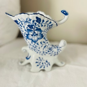 Puede incluir: Un jarrón de cerámica blanca con un diseño floral azul y blanco. El jarrón tiene una forma única con un pico curvo y tres patas.