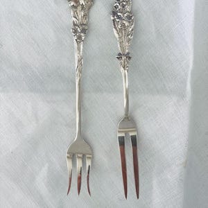 May include: Two silver-toned metal forks with ornate floral handles. The left fork features a three-tine design with a large flower motif at the top. The right fork has two long tines and a handle decorated with a vertical arrangement of small flowers.
