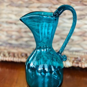 May include: A teal glass jug with a curved handle and a spout. The jug has a rounded base and a slender neck. The glass has a textured appearance, and the jug reflects light. The jug is a decorative item.