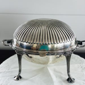 May include: A silver-plated serving dish with a domed, ribbed lid and a smooth, rounded base. The piece features three ornate, curved legs and two side handles. The lid has a circular flat top, and the overall design is metallic and reflective.