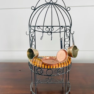 May include: A miniature decorative kitchen scene featuring a black wrought iron structure with a striped tabletop. Small copper and brass pots and pans hang from the structure. A small copper plate sits on the tabletop.
