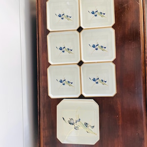 May include: Set of eight square ceramic plates. Each plate features a cream-colored background with a gold trim and a floral design in blue and green. The plates are arranged on a dark wooden surface.
