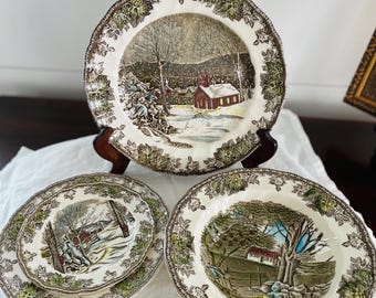 May include: Three vintage ceramic plates with a winter scene featuring a snowy village and a church. The plates have a green and brown floral border.