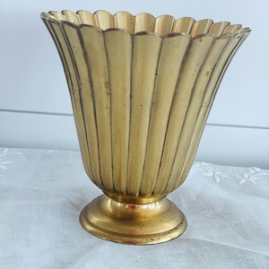 Puede incluir: Un florero de metal color dorado con un borde festoneado y acanalado, que presenta un patrón de costillas verticales a lo largo del cuerpo. El florero tiene una abertura ancha y acampanada que se estrecha hacia una base circular con acabado metálico.