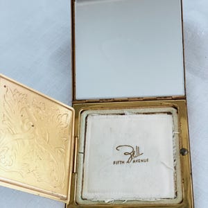 May include: Open vintage gold-toned compact with a mirror and a powder compartment. The compact's lid features an ornate floral design. The powder compartment has a white powder puff with the text "Bell FIFTH AVENUE".