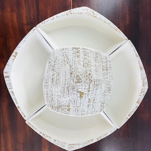 Puede incluir: Una fuente blanca para servir con un centro cuadrado y cuatro compartimentos circundantes. El plato central tiene un acabado texturizado en blanco roto y dorado. Los bordes del plato exterior tienen un ribete dorado envejecido.