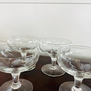 May include: Four clear champagne coupe glasses with etched starburst designs. The glasses have a round bowl shape and sit on a short stem with a circular base. The glasses are arranged on a dark wooden surface.