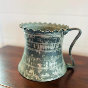 Peut inclure: Une chope en métal antique avec une anse. La chope a une base évasée et un bord festonné. Le métal a un aspect vieilli et patiné avec des bandes horizontales. La poignée est de conception simple et incurvée. La chope est posée sur une surface en bois sombre.