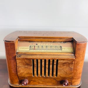 Könnte beinhalten: Ein antikes Bendix-Radio mit einem Holzgehäuse. Das Radio hat ein Einstellrad mit den Zahlen 54, 60, 70, 90, 110, 140 und 170 sowie dem Wort „Police“. Es hat zwei braune Knöpfe und vertikale Lautsprecherschlitze.
