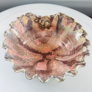 May include: A ceramic bowl with a ruffled edge and a turtle figurine. The bowl has a swirl pattern in shades of brown, pink, and green. The turtle is light brown and is in the centre of the bowl.