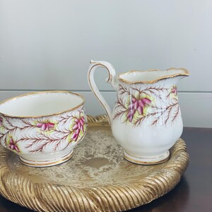 May include: A vintage cream and sugar set with a floral design. The white porcelain pieces feature pink and yellow flowers with gold trim. The set sits on a gold-colored, textured tray, perfect for serving tea or coffee.