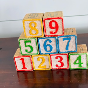 May include: Wooden number blocks with red, yellow, green, and blue numbers. The blocks are stacked on top of each other, displaying the numbers 1 through 9. The blocks are a light brown color.