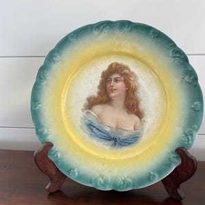 Puede incluir: Un plato de porcelana vintage con un borde verde y amarillo y una imagen pintada de una mujer con el pelo largo y suelto. El plato está sobre un soporte de madera.