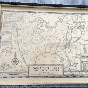 May include: A framed satirical map titled A New Yorker's Idea of THE UNITED STATES OF AMERICA, featuring a distorted geography where New York City dominates the East Coast. The black and white print includes text blocks, a compass rose, and illustrations of city landmarks, all enclosed in a decorative black and gold frame.