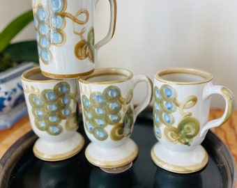 May include: Set of four ceramic mugs with a white base, featuring a grape design in blue, green, and brown. The mugs have a pedestal base and a handle. The mugs are on a black tray.