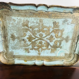 May include: An antique rectangular serving tray with a distressed finish. The tray features a light blue center with gold-colored ornate floral designs and a gold-colored scalloped edge. The tray is a decorative item.