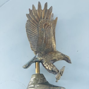 Pode incluir: Uma escultura de águia em bronze com as asas abertas, segurando um pequeno objeto nas garras. A águia é montada em uma pequena base texturizada. A escultura apresenta penas detalhadas e um acabamento polido.