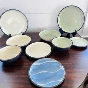 May include: A collection of ceramic dinnerware, including plates and bowls, in various sizes and colors. The plates have a cream or light green base with a dark blue rim. One plate features a blue wave design.