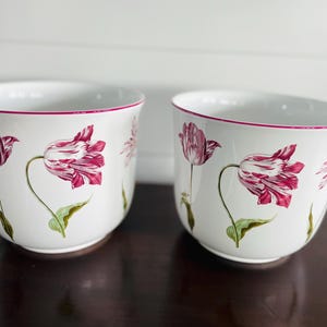 May include: Two white ceramic planters with a floral tulip design. The tulips are a mix of white and deep pink, with green stems and leaves. The rims are trimmed with a thin pink line. These planters are ideal for indoor or outdoor use.