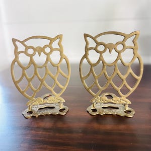 May include: Two brass owl-shaped bookends. The bookends have a cut-out design with a detailed owl face and body. The base of each bookend has a decorative floral pattern. The bookends are displayed on a dark brown wooden surface.