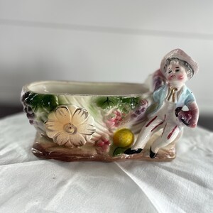 May include: A ceramic planter featuring a sculpted figure of a person wearing a hat and light blue jacket, seated on the edge. The vessel is decorated with relief carvings of a large flower, green leaves, clusters of purple grapes, and a yellow fruit.
