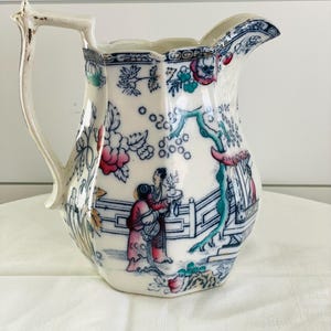 May include: Antique white ceramic pitcher with a detailed Asian-inspired design. The pitcher features a floral and landscape pattern in shades of blue, pink, and green. The handle has a unique, ornate shape.