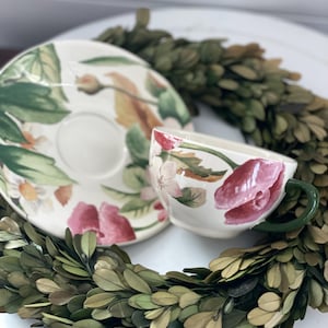 May include: A floral teacup and saucer set with a green handle and a matching floral design. The cup and saucer are surrounded by a green wreath. The floral design includes pink and white flowers with green leaves.