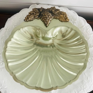 May include: A scalloped shell-shaped dish with a pale green metallic finish. The top edge features a decorative bronze-toned metal accent shaped like a cluster of grapes with detailed leaves, providing a contrast to the smooth, ribbed surface.