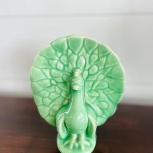Puede incluir: Figura de pavo real de cerámica verde. El pavo real tiene una cola detallada y se apoya sobre una pequeña base. La figura es un objeto decorativo, probablemente para exhibición, en un verde brillante.