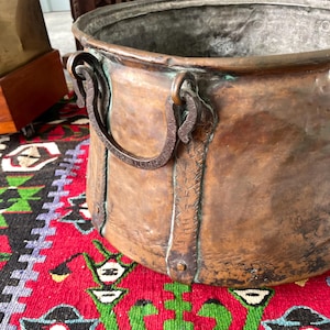 May include: A large, round, hammered copper pot featuring a wide opening and a dark, weathered patina. The vessel includes two heavy, curved iron handles attached to the sides with vertical metal straps that show etched, cross-hatched detailing.