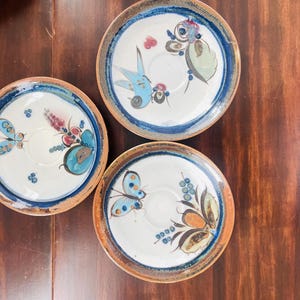 May include: Three round ceramic saucers with a white center and a brown rim. Each saucer features a hand-painted design of a blue butterfly and floral motifs in shades of blue, orange, and green. The edges are accented with a dark blue band.