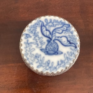May include: A round, white and blue porcelain trinket box lid with a silver-colored filigree edge. The lid features a blue floral design in the center. The background is a dark wood surface.