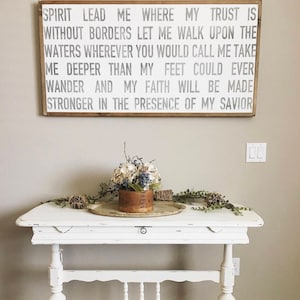 Puede incluir: Un letrero de madera blanco con el texto "SPIRIT LEAD ME WHERE MY TRUST IS WITHOUT BORDERS LET ME WALK UPON THE WATERS WHEREVER YOU WOULD CALL ME TAKE ME DEEPER THAN MY FEET COULD EVER WANDER AND MY FAITH WILL BE MADE STRONGER IN THE PRESENCE OF MY SAVIOR". El letrero está colgado en una pared beige claro sobre una mesa blanca desgastada con un cajón. Un pequeño ramo de flores en una maceta marrón está sobre la mesa.