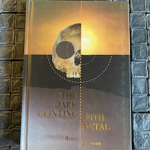 May include: A book cover with a dark brown background and a skull with a black eye socket in the center. The title "The Dark Glinting With Metal" is written in gold lettering. The author's name, "Brian Evenson", is written in smaller gold lettering below the title.