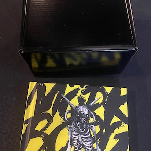 Può includere: Una scatola di cartone nera con un design grafico giallo e nero sul fondo. La scatola è appoggiata su un libro con una copertina gialla e un'illustrazione in bianco e nero di uno scheletro con le corna. Il titolo del libro è "WIRK ROAR" e il testo sulla copertina recita "Text and game design Pelle Nilsson Graphic design and art Johan Nohr Dead Noods".