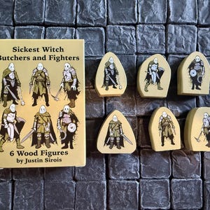 Op de afbeelding: Zes houten figuren van personages uit het spel Sickest Witch Butcher's and Fighters. De figuren zijn geschilderd in tinten bruin en wit en staan op een grijs stenen oppervlak.