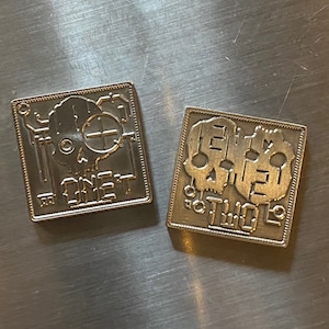 May include: Two silver metal pins with a skull design. The first pin says "ONE" and the second pin says "TWO".